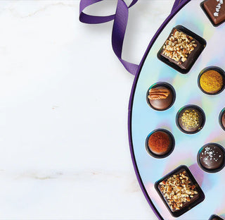 Vosges Chocolate Ensemble circular candy box sits open displaying a collection of bonbons, truffles, caramel covered marshmallows and toffee ornamented with a purple ribbon on a white background.