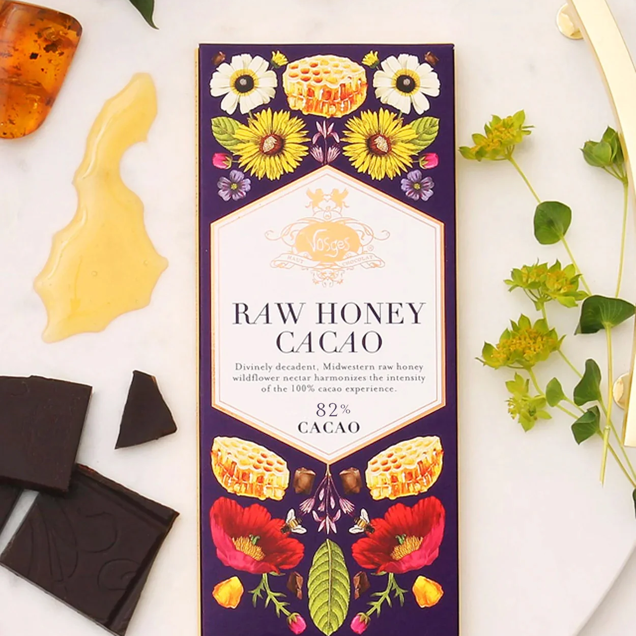 A 3 ounce Vosges Raw Honey Cacao bar besides a dap of raw honey pieces of dark chocolate and some flowers